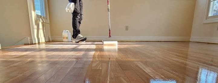 A coating being rolled onto a timber floor on an extension pole, the wet edge still reflecting the windows